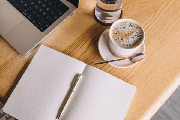 Laptop sitting on table with notebook, pen, glass of water, and cup of coffee