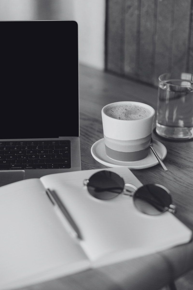 laptop on table with notebook and sunglasses sitting on top of it next to a cup of coffee and cup of water